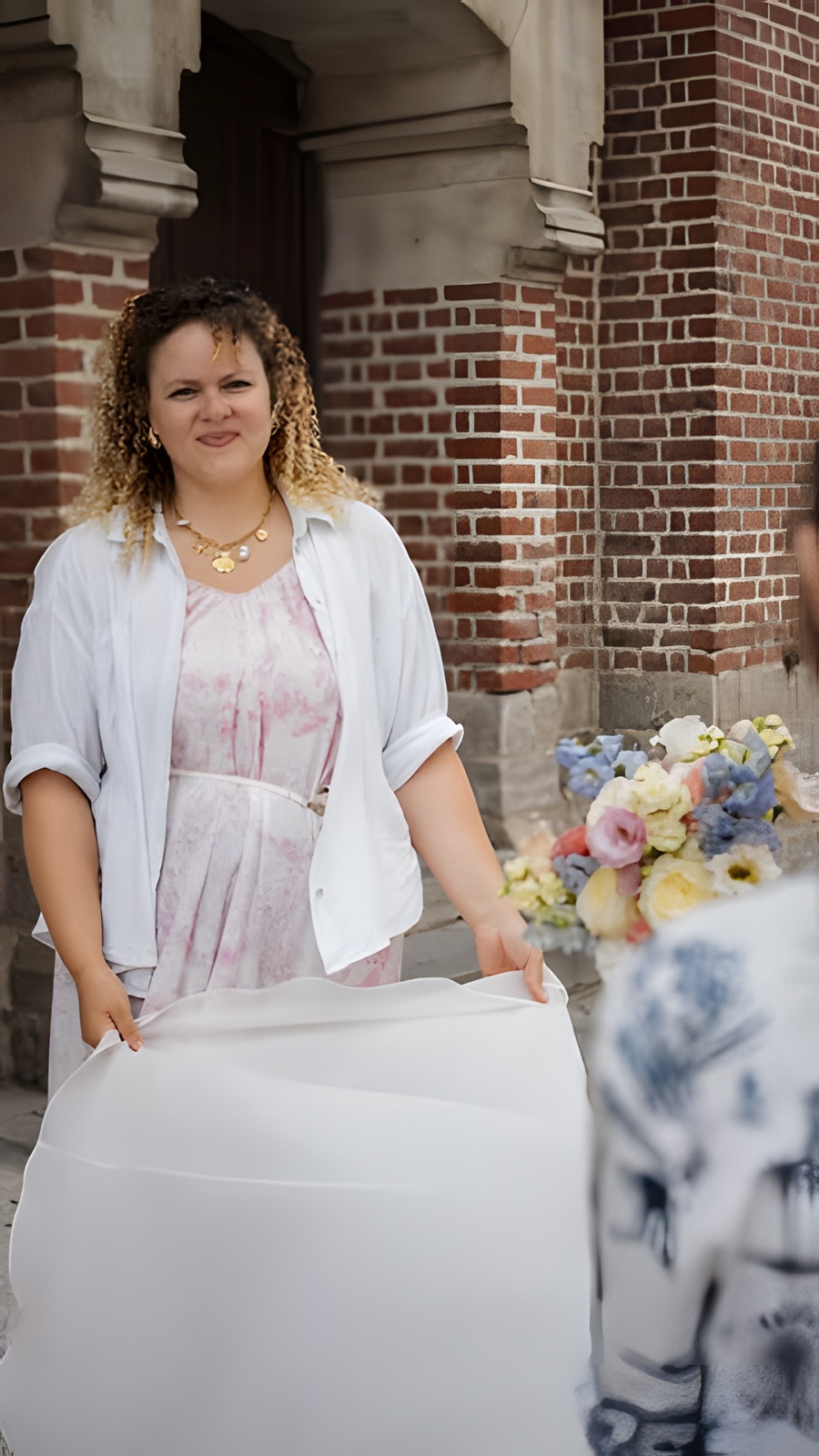 Oriane, en robe à fleurs et chemise blanche, tient la traîne de la mariée devant l'église.